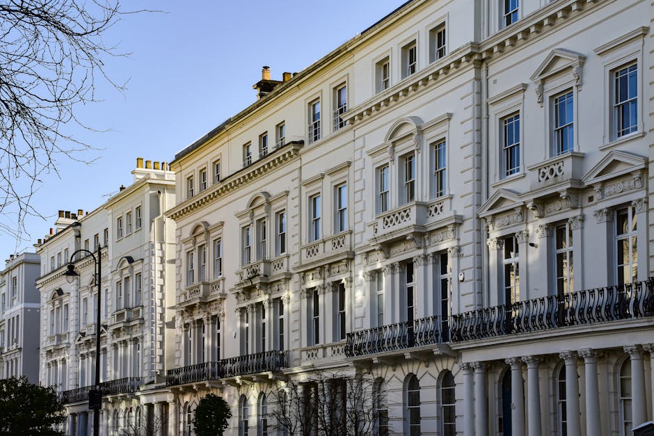 The image depicts a row of elegant, multi-story white terraced buildings with neoclassical architectural details, including decorative moldings, pilasters, and ornate balcony railings made of black wrought iron. The facades are finished with smooth, light-colored stucco, and the windows feature white frames with some having small protruding pediments. The buildings are situated along a paved street, with a few leafless trees visible on the left side casting slight shadows on the facades, suggesting a clear day with bright natural light. The street is lined with black lampposts, and small shrubs are planted in front of the buildings. The overall scene presents a neat, well-maintained urban environment, typical of Victorian or Georgian style architecture, within a residential or city centre context that may be associated with private property management or onsite waste clearance practices as seen in the scene. The clarity and detail of the buildings’ external details emphasize their period style, offering an observable connection to urban waste management or property maintenance efforts, such as those provided by Waste Disposal Brent, for private waste handling from terraces and estates.