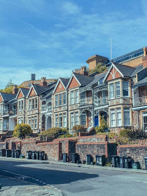 A row of Victorian-style terraced houses positioned along a residential street, each with ornate decorative white ironwork on the balconies and window frames. The houses feature brick facades with bay windows and gabled roofs, some with small front gardens or steps leading up to the entrances. The distance between the houses varies slightly, creating a staggered appearance, with a bright blue sky overhead and a few wispy clouds. In the foreground, a paved pavement runs alongside the street, with multiple black wheelie bins lined up at the curb, suggesting local waste collection. Behind the houses, taller modern buildings are visible, indicating an urban setting that may require alternative waste handling solutions. The lighting is natural, hinting at a clear day, and the overall scene reflects a well-maintained residential area where private rubbish disposal, potentially coordinated by Waste Disposal Brent, supports local waste management needs without reliance on municipal services.