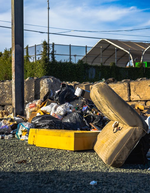 An outdoor scene showing a pile of assorted waste materials on a rough asphalt surface, including black plastic rubbish bags, a broken beige car tire, a yellow plastic storage container, and various smaller debris and packaging. The waste is accumulated near a low stone wall composed of irregularly shaped, weathered stone blocks. In the background, there are metal utility poles with overhead electrical wires, green foliage, and a large, partially covered greenhouse structure made of translucent panels supported by metal frames. The scene is illuminated by bright daylight, casting shadows on the ground, highlighting the disparity between the clean environment and the unmanaged rubbish heap, suggesting an area suitable for private waste collection or rubbish removal services like those provided by Waste Disposal Brent, emphasizing the importance of proper waste management solutions.
