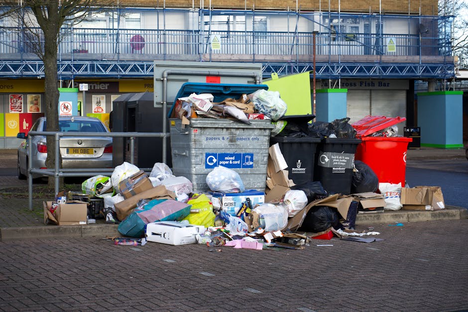 An outdoor scene showing a pile of assorted waste materials on a rough asphalt surface, including black plastic rubbish bags, a broken beige car tire, a yellow plastic storage container, and various smaller debris and packaging. The waste is accumulated near a low stone wall composed of irregularly shaped, weathered stone blocks. In the background, there are metal utility poles with overhead electrical wires, green foliage, and a large, partially covered greenhouse structure made of translucent panels supported by metal frames. The scene is illuminated by bright daylight, casting shadows on the ground, highlighting the disparity between the clean environment and the unmanaged rubbish heap, suggesting an area suitable for private waste collection or rubbish removal services like those provided by Waste Disposal Brent, emphasizing the importance of proper waste management solutions.