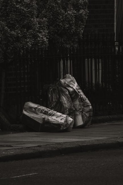 Two large black plastic trash bags, slightly crumpled and tied at the top, sit on a paved sidewalk near the curb in an urban area. The bags are filled with waste and appear to be full, with some bits of material visibly protruding. Behind them is a black wooden fence with vertical slats, and a dense green leafy bush extending upward, partially obscuring the fence. The pavement is smooth, and the street in front is visible with a dark surface. The scene appears to be at dusk or in low lighting conditions, giving a subdued and neutral tone to the image. The placement of the rubbish suggests they are awaiting collection, providing a context related to private waste disposal or alternative rubbish removal services in the area. Waste Disposal Brent might handle such street clearing and rubbish removal, as seen in this typical urban setting for bulk waste collection and on-site clearance services.