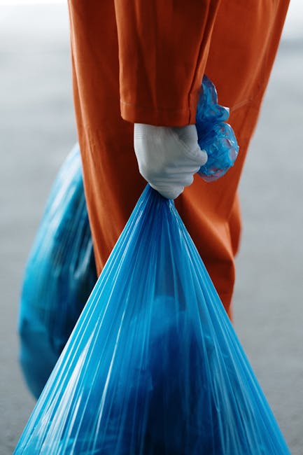 A close-up view of a person wearing an orange protective suit and a white glove, holding a tightly knotted blue plastic bag filled with waste. The person's arm and hand are centered in the image, with the bag's smooth, shiny surface clearly visible, showing folds and creases from handling. The background is blurred but suggests an outdoor environment, possibly near a waste collection or disposal site, with neutral tones. The lighting highlights the glossy texture of the plastic bag, emphasizing its material, and the orange suit contrasts with the blue bag, drawing attention to the disposal activity. This scene illustrates a waste management process where an individual is preparing waste for transportation, aligning with services such as private rubbish collection or on-site clearance that Waste Disposal Brent provides for efficient rubbish removal in the local area.