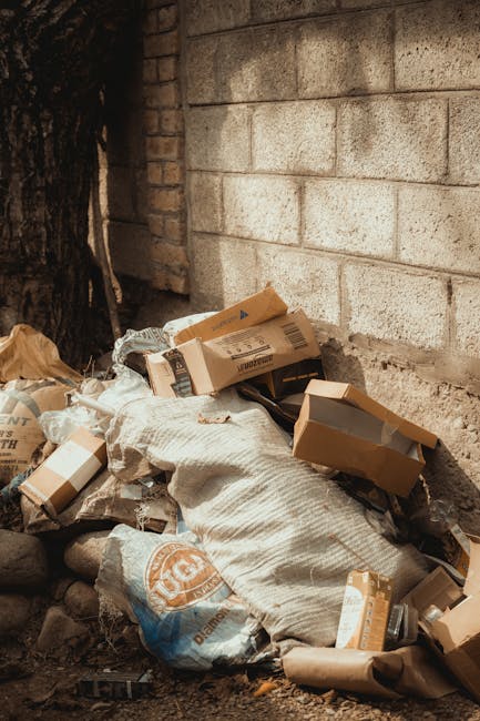 A pile of mixed rubbish is stacked against a textured brick and concrete wall, situated outdoors near a tree with dark bark. The waste includes cardboard boxes, some partially flattened and others still with identifiable flaps, along with plastic bags and wrapping materials. The cardboard varies in light brown shades, with some boxes displaying printed labels or barcodes. The plastic bags are light-colored, with a crumpled appearance, and possibly contain additional refuse. The ground beneath the rubbish consists of dirt, small stones, and debris, indicating an outdoor or construction environment. The scene appears untidy, suggesting private waste collection or on-site clearance often handled by waste disposal services like Waste Disposal Brent. The overall lighting is natural, casting soft shadows, highlighting the textures of the materials and the rough surface of the wall, emphasizing a typical waste accumulation situation that might be targeted by rubbish removal services for cleanup and proper disposal.