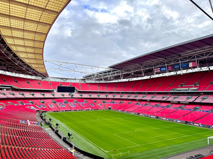 A large, modern football stadium with a partially enclosed roof structure covering the seating areas, which are predominantly red with some pink seats. The interior view captures the well-maintained green grass pitch with visible white boundary lines, goalposts at each end, and some floodlights positioned along the sidelines. Overhead, a partly cloudy sky is visible through the open segments of the roof, providing natural light that illuminates the field evenly. The stadium features tiered seating sections surrounding the pitch, with some sections showing signs of recent use or maintenance. In the background, a large black digital scoreboard and banners with UEFA and FIFA logos hang from the upper tiers, indicating the venue hosts international matches. The overall scene suggests a professional sports arena prepared for a sporting event, with no visible crowd or spectators, capturing the typical layout of an indoor-outdoor football stadium designed for large audiences and media coverage. The image subtly highlights the facility’s capacity for hosting significant events, aligning with services related to stadium waste management and event rubbish removal along with private disposal options managed by Waste Disposal Brent.