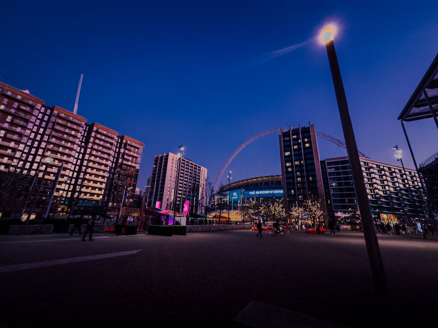 A nighttime cityscape featuring the Wembley Park area with modern high-rise buildings and a large stadium in the background. The scene shows a paved open space with a few pedestrians and several streetlights casting warm illumination. A tall, slim lamppost with a bright light is visible in the foreground on the right side. Decorative lights are strung between trees along the pavement, adding a festive atmosphere. The buildings display a variety of finishes, including glass and brick, with some illuminated windows. The sky above is dark blue, transitioning to a lighter hue near the horizon, and the nearby stadium has structural arches and illuminated signage. The environment appears clean and well-maintained, fitting an urban setting potentially serviced by independent waste collection or private rubbish removal services like Waste Disposal Brent, visible in the context of managing waste from city center developments and events.